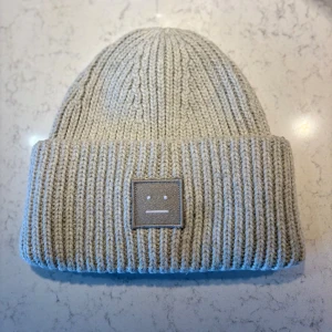 Grå Acne Studios Mössa - Snygg beige ribbstickad mössa från Acne Studios med klassisk uppvikt kant och det ikoniska Face-logotypen framtill. Mjuk och varm, perfekt för kyliga dagar. Märkt med Acne Studios på insidan. Stilren och enkel att matcha med din garderob.