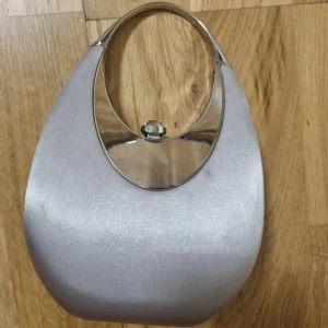 Silver axelväska med metallhandtag - Snygg axelväska i silver med ett unikt, ovalt metallhandtag och blank finish. Väskan har en modern, rundad form och stängs med ett vridlås i metall. Perfekt för dig som gillar stilrena och trendiga accessoarer.