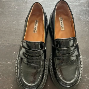 Svarta chunky loafers från NA-KD - Säljer ett par svarta loafers från NA-KD med chunky sula och glansig finish. Skorna har klassisk penny loafer-design och grovt mönstrad yttersula för extra grepp. Perfekta för dig som vill ha en edgy men stilren look.