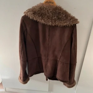 Brun teddyjacka med fuskpäls - Mysig brun teddyjacka med fluffig fuskpäls på krage, ärmslut och insida. Jackan har en snygg retrovibe, knäpps med knappar framtill och har en loose passform. Perfekt för kalla dagar när du vill vara både varm och stilren. Skriv vid frågor💕