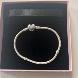 Pandora armband med hjärtformad lås - Snyggt silverfärgat armband från Pandora med ett hjärtformat lås. Armbandet har en tvinnad design och låset är dekorerat med Pandora-loggan. Perfekt för att kombinera med dina favoritberlocker. Helt nytt, enbart testat men tyvärr för stort! 