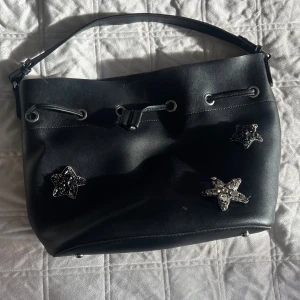 Svart axelväska med stjärndetaljer - Svart axelväska från Lantadeli Paris i skinn med coola glittriga stjärnor framtill. Väskan har snörning upptill och ett rymligt innerfack med dragkedja. Perfekt storlek för allt du behöver och snygg att bära över axeln.