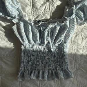 Blommig volangtopp från Gina Tricot - Supersöt ljusblå blommig topp från Gina Tricot med smockad midja, volangkant nertill och knyt framtill. Toppen har offshoulder-modell och är perfekt för vår och sommar. Tunt och luftigt material som känns skönt mot huden.