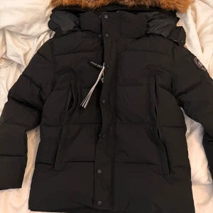  Canada Goose Wyndham Parka black label - Säljer en svart dunjacka från Canada Goose i storlek XS. Jackan har en fluffig huva med brun päls, två stora fickor med dragkedja och snygga detaljer som logopatch på ärmen. Perfekt för kalla vinterdagar och har en klassisk, stilren look.