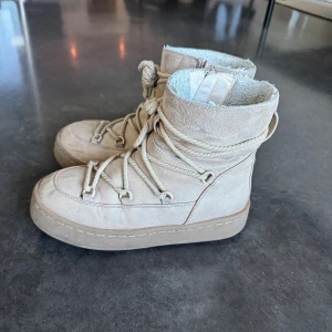 Varmfodrade boots med snörning - Säljer ett par beiga varmfodrade boots med snörning och mjukt foder. Skorna har en platt sula och är tillverkade i mocka med detaljer i rep och metallöglor. Perfekta för kalla dagar när du vill hålla stilen och värmen. Nypris ca 500 kr.