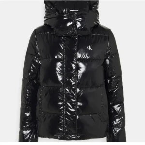 Svart glansig pufferjacka Calvin Klein - Säljer en svart, glansig pufferjacka från Calvin Klein med CK-logga på bröstet. Jackan har hög krage, huva och dragkedja framtill. den är lite mindre i storleken . 
