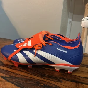 Adidas Predator blå/orange fotbollsskor - Säljer ett par Adidas Predator fotbollsskor i storlek 42. Skorna är blå med vita och orange detaljer, har snörning och en låg profil. Snygg design med klassiska Adidas-ränder och Predator-tryck på sidan. Perfekta för dig som vill sticka ut på planen.