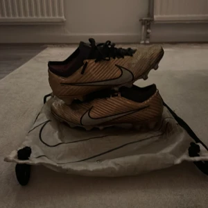 Nike Zoom fotbollsskor guld/svart - Säljer ett par Nike Zoom fotbollsskor i guld med svarta detaljer och snörning. Skorna har en platt sula med fasta dobbar för bra grepp på planen. Ovandelen är tillverkad i syntetmaterial med textur för bättre bollkontroll. Snygg och modern design som sticker ut.
