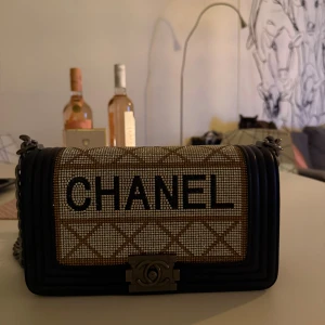  axelväska med paljetter - Snygg axelväska  med glittriga paljetter och tydligt logotyp framtill. Väskan har ett rutigt mönster i beige och guldtoner, svart kant och grov kedja som axelrem. Perfekt för dig som vill sticka ut med en lyxig accessoar.