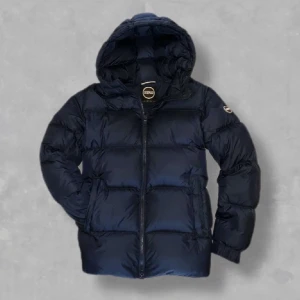 Mörkblå pufferjacka från Colmar - Säljer en mörkblå pufferjacka från Colmar med huva och dragkedja framtill. Jackan har en quiltad design och två sidofickor. Perfekt för kalla dagar och riktigt snygg till streetwear-stilen.