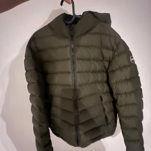 Olivgrön Colmar dunjacka med huva - Snygg olivgrön dunjacka från Colmar med huva och dragkedja framtill. Jackan har quiltad design, två fickor med dragkedja och en rund Colmar-logga på ärmen. Perfekt för kyliga dagar och riktigt skön att bära. Obs! Köpte jackan för 5000 men pga de pyttesmå hålen på armen så har jag satt ned priset så pass mycket!