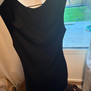 Svart ärmlös kortklänning - Snygg svart klänning med ärmlös design, rundad hals och öppenrygg. Modellen är figurnära och har en klassisk, enkel stil som passar dig som gillar minimalistiskt mode. Klänningen är kort och har en slät yta utan mönster eller detaljer.