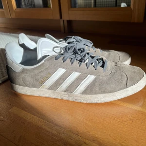 Adidas Gazelle grå sneakers strl 46 - Säljer ett par klassiska Adidas Gazelle sneakers i grå mocka med vita detaljer och de ikoniska tre ränderna på sidan. Skorna har snörning och platt sula, samt diskret guldfärgad Gazelle-text på sidan. Perfekta för dig som gillar stilrena och tidlösa sneakers.