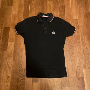 Svart Moncler pikétröja med röd/vit detalj - Svart pikétröja från Moncler med klassisk krage och knappar framtill. Tröjan har Moncler-logga på bröstet och snygga rödvita ränder på kragen och ärmsluten. Tillverkad i bomull för skön känsla och stilren look.