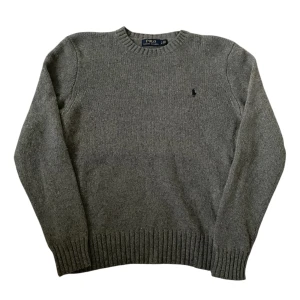 Grå stickad tröja från Polo Ralph Lauren | L - Klassisk grå stickad tröja från Polo Ralph Lauren med rund halsringning och diskret broderad logga på bröstet. Tröjan har ribbade muddar vid ärmslut och nederkant, perfekt för lager-på-lager under kyliga dagar.