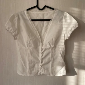 Vit blus med spets och knappar - Säljer en söt vit blus med korta puffärmar, djup v-ringning och spetskant längs halsen. Blusen har dekorativa knappar framtill och fina sömmar som ger en feminin siluett. Perfekt för dig som gillar romantisk och stilren look.