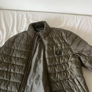 True Religion pufferjacka blå & grön - Två snygga pufferjackor från True Religion, en i blått och en i olivgrönt. Jackorna har quiltad design, hög krage och dragkedja framtill. True Religion-loggan är tryckt på bröstet. Perfekt för dig som gillar streetwear och vill ha en varm jacka med cool look.