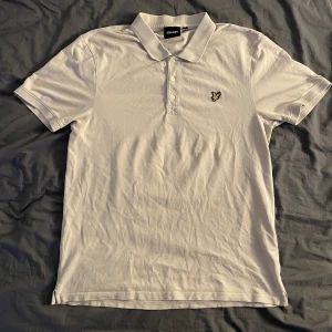 Beige piké från Lyle & Scott i XL - Beige pikétröja från Lyle & Scott med klassisk krage och knappar framtill. Tröjan har det ikoniska gula fågel-loggan broderad på bröstet. Tillverkad i bomull och har korta ärmar. Perfekt för en clean och stilren look.