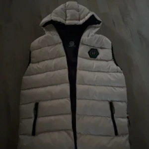 Vit dunväst från Philipp Plein - Snygg dunväst från Philipp Plein i ljusvit färg med svart insida och detaljer. Västen har huva, två fickor med dragkedja och ett coolt PP-emblem på bröstet. Stora svarta dragkedjor och quiltad design ger en modern vibe. Perfekt för dig som vill sticka ut.