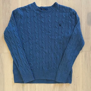 Blå kabelstickad tröja Polo Ralph Lauren - Säljer en blå kabelstickad tröja från Polo Ralph Lauren i storlek S. Tröjan har rund hals, ribbade muddar och det klassiska broderade logot på bröstet. Perfekt för dig som gillar stilrena och tidlösa plagg med snygg struktur.
