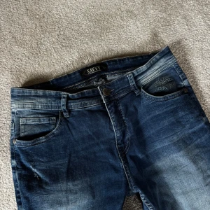 Mörkblå jeans från Savvy - Snygga mörkblå jeans från Savvy med klassisk femficksdesign och coola slitningar på låren. Jeansen har rak passform och normal midja, tillverkade i mjukt denimtyg som sitter skönt hela dagen. Perfekt för dig som gillar en avslappnad stil.