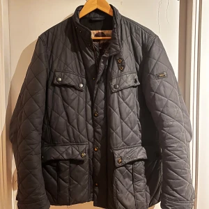 Barbour Jacka grå - Quiltad grå jacka från Barbour i storlek M. Jackan har ståkrage med tryckknappar, flera fickor med lock och knapp, samt flera snygga detaljer. Jackan har en klassisk, normal passform och är perfekt för hösten. 