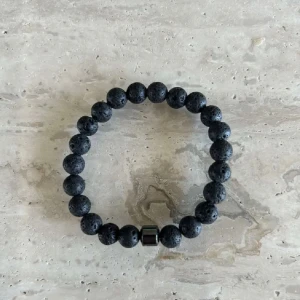 Svart armband  - Snyggt svart armband med runda lavastenpärlor och en metallisk detalj. Perfekt accessoar för dig som gillar en stilren och cool look. Elastisk tråd gör det enkelt att ta på och av. Passar både killar och tjejer.