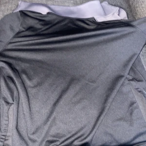 Svart och grå träningsjacka Under Armour - Säljer en svart och grå träningsjacka från Under  med hel dragkedja och hög krage. Jackan har en sportig look och är tillverkad i ett mjukt syntetmaterial som passar perfekt för träning. Stort Under Armour-logga på bröstet.