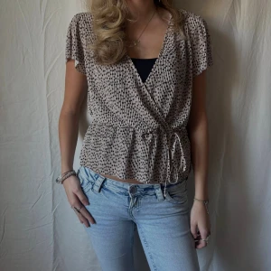 Prickig omlottblus - Beige omlottblus / peplumtopp med svart prickigt mönster. Blusen har korta ärmar, knyt i midjan och v-ringning. Snygg till både vardag och fest.