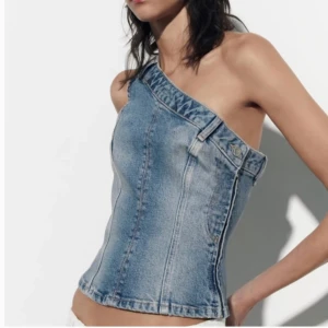 Jeans one shoulder topp från Zara - Säljer en ljusblå jeans one shoulder topp från Zara. Toppen har en asymmetrisk skärning, bälteshällor och dragkedja i sidan för enkel påklädning. Snygg  modell i klassiskt denimtyg som ger en cool och trendig look.