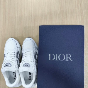 Vita Dior sneakers med grå detaljer - Säljer ett par fräscha vita Dior sneakers med grå och svarta detaljer. Skorna har tydlig CD-logga på sidan och baksidan, samt snygg snörning. Ovandelen är i mesh och skinn för en modern look. Perfekta för dig som vill sticka ut med exklusiva sneakers.