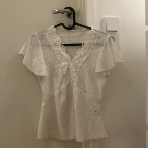 Vit spetsblus med volangärm - Superfin vit blus med broderade detaljer och spetskantad v-ringning. Blusen har korta volangärmar och lätt utsvängd nederdel. 