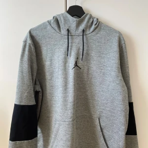 Grå Jordan hoodie med svarta detaljer - Snygg grå hoodie från Jordan med svart logga på bröstet och svarta partier på ärmarna. Tröjan har huva med snörning och en stor magficka. Perfekt för dig som gillar sportig och avslappnad stil.