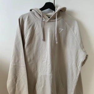 Beige hoodie från Gymshark - Säljer en stilren beige hoodie från Gymshark med huva och dragsnören. Tröjan har en klassisk känguruficka framtill och diskret logga på bröstet. Perfekt för chill dagar eller till gymmet.