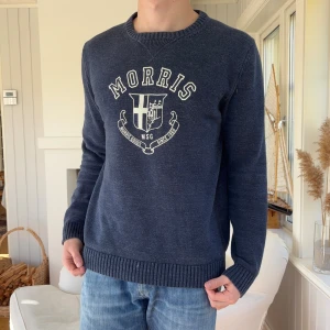 Mörkblå stickad tröja från Morris - Säljer en mörkblå stickad tröja från Morris med stort tryck och emblem på bröstet. Tröjan har rund halsringning, ribbade muddar och en klassisk, avslappnad passform. Perfekt för dig som gillar preppy stil och snygga detaljer.