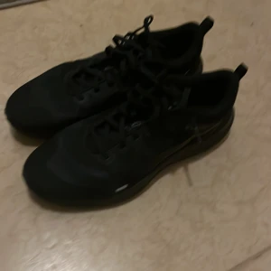 Svarta Nike sneakers med snörning - Säljer ett par helsvarta Nike sneakers med klassisk snörning och rund tå. Skorna har en sportig vibe med diskret Nike-logga på sidan och dragflikar bak för enkel påtagning. Ovandelen är i syntetmaterial och sulan är platt och bekväm.