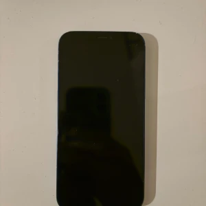 iPhone 12 Blå - Säljer en blå iPhone 12. Telefonen har sprickor och repor på baksidan samt slitage på kanterna. Batterihälsan är 76% och behöver bytas för optimal prestanda. Skärmen ser hel ut. Perfekt för dig som vill ha en prisvärd iPhone med dubbla kameror.