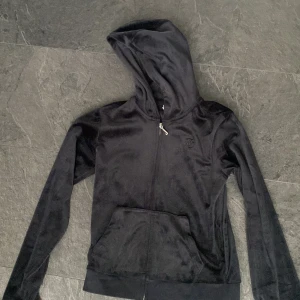 Svart velour hoodie från Juicy Couture - Mjuk och skön svart hoodie i velour från Juicy Couture. Klassisk modell med huva, dragkedja framtill och känguruficka. Liten broderad logga på bröstet och ribbade muddar vid ärmslut och nederkant. Perfekt för chill dagar.