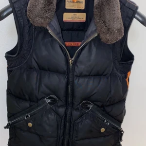 Svart dunväst från Parajumpers - Svart dunväst från Parajumpers med brun fuskpälskrage, två snedställda fickor med dragkedja och tryckknapp. Västen har quiltad design och markerad krage, perfekt för kalla dagar. Slim fit-modell med coola detaljer och orange inslag på sidan.