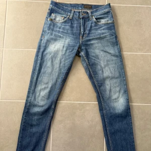 Tiger of Sweden blå jeans Evolve 28/32 - Snygga blå jeans från Tiger of Sweden i modellen Evolve. Jeansen har klassisk femficksdesign, orangea kontrastsömmar och en något tvättad look. Tillverkade i Italien av jeansmaterial och har en rak passform som sitter snyggt på benen.