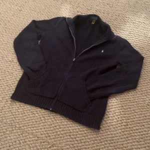 Svart kofta från Polo Ralph Lauren - Svart kofta från Polo Ralph Lauren med dragkedja framtill och två fickor. Klassisk design med ribbade muddar och diskret logga på bröstet. Perfekt för dig som gillar stilrena och tidlösa plagg. Materialet är mjuk bomull och koftan har långärmad passform.