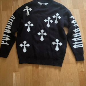 Svart stickad tröja med vita kors - Cool svart stickad tröja med vita kors och grafiska detaljer på ärmarna. Tröjan har rund hals och långärmad passform, perfekt för dig som gillar edgy och unik stil. Materialet är mjukt och håller dig varm under kyliga dagar. För kvinnor passar det mer som S/M