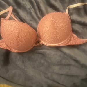 Rosa spetsig bh Push up - Säljer en snygg rosa bh med spetsdetaljer och vadderade kupor. BH:n har tunna axelband och en liten guldfärgad detalj framtill. Perfekt för dig som gillar romantisk och feminin stil.