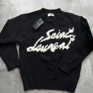 Svart stickad Saint Laurent tröja - Svart stickad tröja från Saint Laurent med vit, fluffig logotyp framtill. Tröjan har rund halsringning och långa ärmar. Perfekt för dig som vill ha en statement piece med lyxig känsla och cool design.