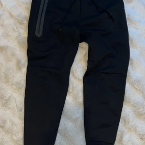 Svarta nike tech byxor ny modell - Svarta Nike tech byxor från Nike. Köptes för 1250kr från JD men max använd 1-2. Skulle säga att den är i nyskick eftersom jag bara hade på mig den några timmar.