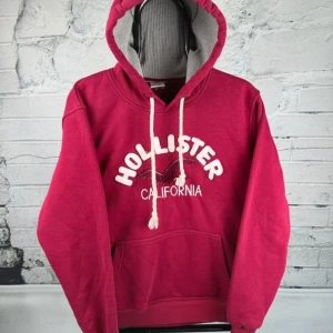 Röd hoodie från Hollister med tryck - Snygg röd hoodie från Hollister med broderat vitt logotyp-tryck och fågel på bröstet. Tröjan har huva med vita snören, känguruficka och ribbade muddar. Perfekt för chill dagar och streetstyle. Mjuk insida och klassisk passform.