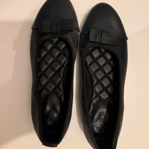 Svarta ballerinaskor med rosett Jenny Fairy - Snygga svarta ballerinaskor från Jenny Fairy med rund tå och dekorativ rosett framtill. Skorna har quiltad innersula för extra komfort och en platt sula. Yttermaterialet har en matt finish och insidan är mjukt vadderad.