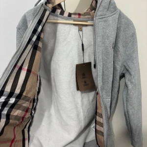 Grå Burberry kofta - Snygg grå Burberry med klassiskt rutigt foder i huvan och insidan. Perfekt för dig som gillar stilrena och exklusiva plagg med en twist.