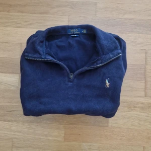 Mörkblå half zip tröja Polo Ralph Lauren - Mörkblå långärmad tröja från Polo Ralph Lauren med broderad logga på bröstet. Tröjan har en klassisk half zip-dragkedja och ribbade muddar. Tillverkad i bomull och passar perfekt för lager-på-lager under höst och vinter.