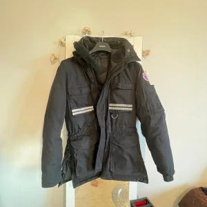 Svart parkajacka från First Slope - Svart parkajacka från First Slope med huva, reflexdetaljer fram och bak samt flera stora fickor. Jackan har broderad patch på ärmen och mjukt foder på insidan. Perfekt för kalla dagar när du vill hålla stilen och värmen.model är stor